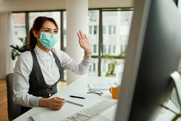 Happy businesswoman wearing face mask and waving while having video chat over computer in the office.