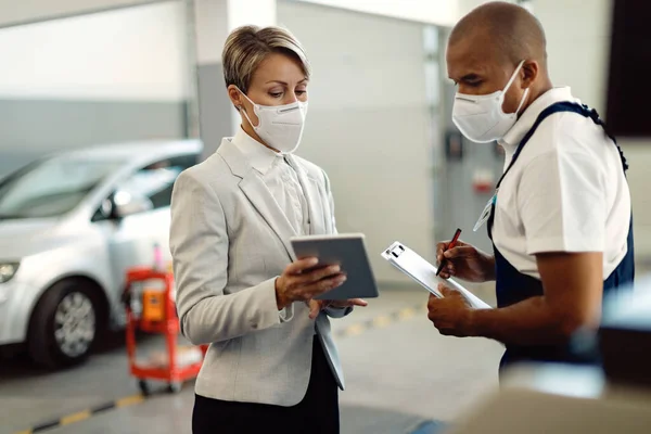 Black auto repairman and businesswoman using digital tablet at car service workshop and wearing face masks due to coronavirus pandemic. Focus is on woman.