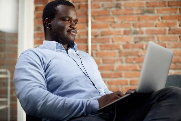 Smiling black businessman with in-ear headphones surfing he net on laptop in the office.