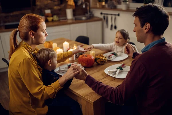 Parents and their kids holding hands and praying before Thanksgiving dinner in dining room.