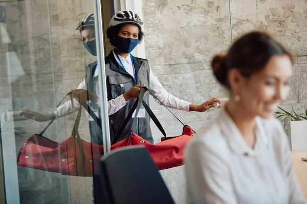 Black food deliver with face mask carrying thermal backpack while delivering lunch at business office.