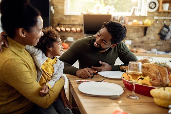 Happy black parents and their small daughter having Thanksgiving lunch and communicating at dining table.