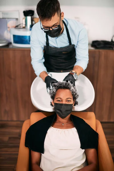 Black woman and her hairdresser wearing protective face masks during hair wash at the salon.