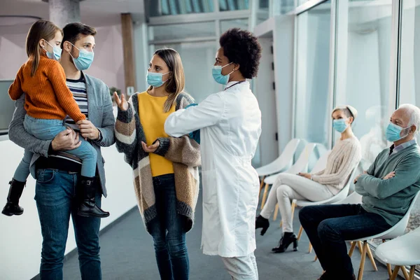 Parents with daughter communicating with black female doctor while wearing fsce masks in waiting room at the hospital.