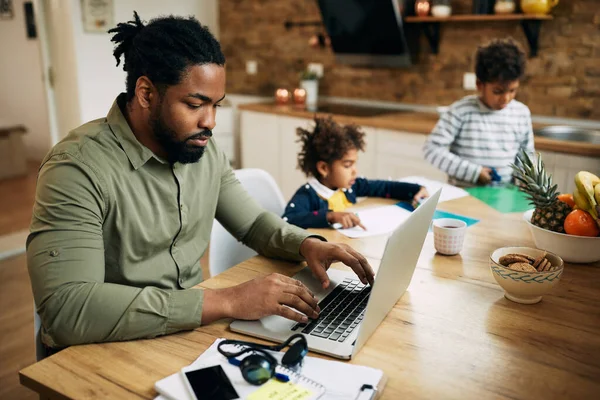 Black stay at home father working on a computer while his children are playing in the background.