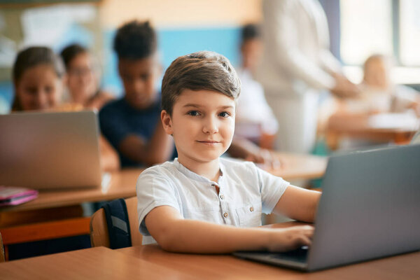 Милый школьник учится за ноутбуком во время занятий в классе и смотрит в камеру. 