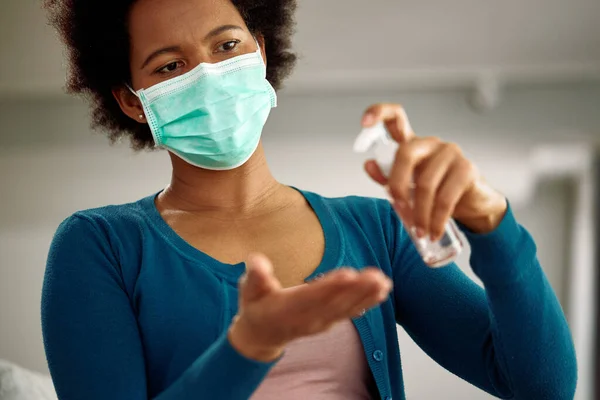 Black woman with face mask using hand sanitizer at home during virus pandemic.