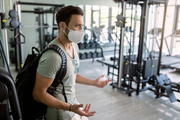 Athletic man with protective face mask feeling confused while standing in empty gym. 