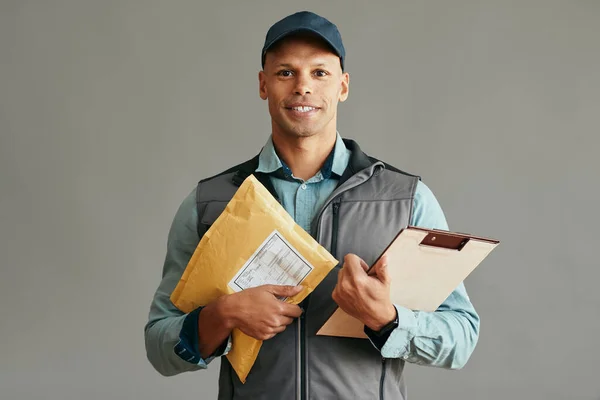 Happy African American postal worker holding package and clipboard against gray background and looking at camera. Copy space.