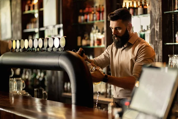 Young bartender working at bar counter and pouring beer in a glass.