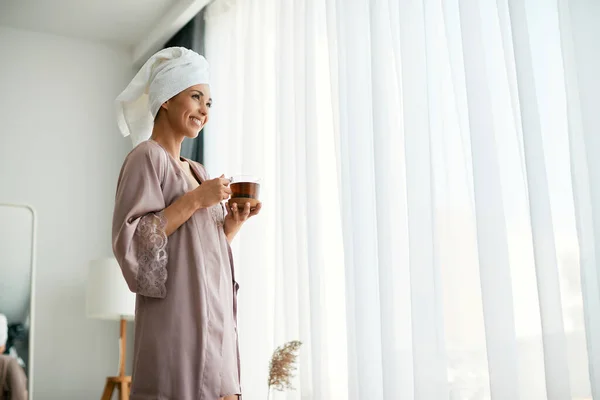 Young happy woman in nightgown drinking tea and looking through the window at home. Copy space.