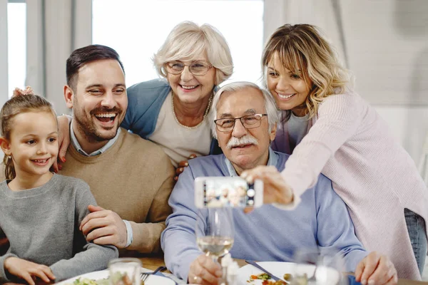 Happy extended family using smart phone taking selfie with smart phone in dining room.