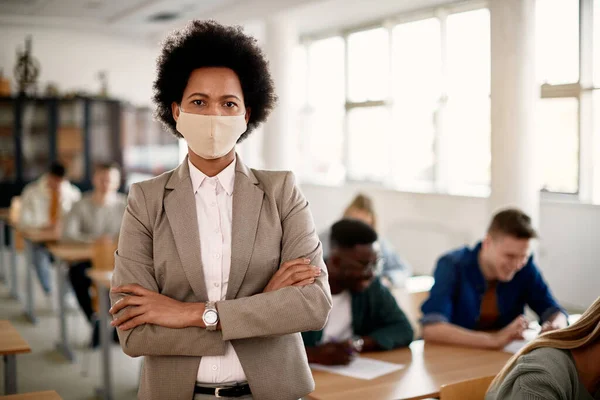 Black female professor with protective face mask standing with crossed arms at lecture hall and looking at camera.