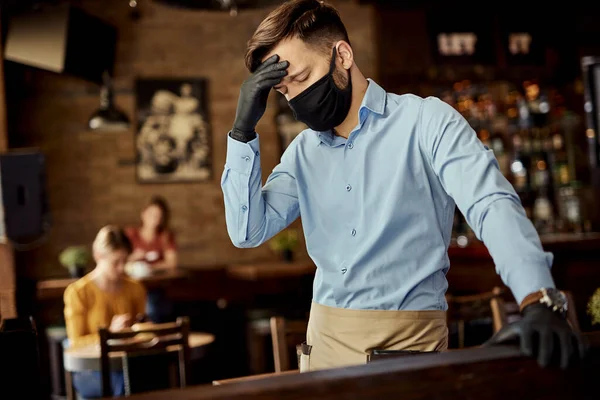 Young waiter with protective mask on is face feeling tired and holding his head in pain while working in a cafe.
