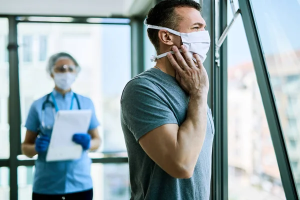 Pensive man with face mask feeling COVID-19 symptoms and looking through the window while being at medical clinic. Nurse is standing in the background.