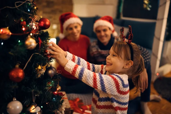 Happy Little Girl Putting Ornaments Christmas Tree Home Her Parents — Zdjęcie stockowe