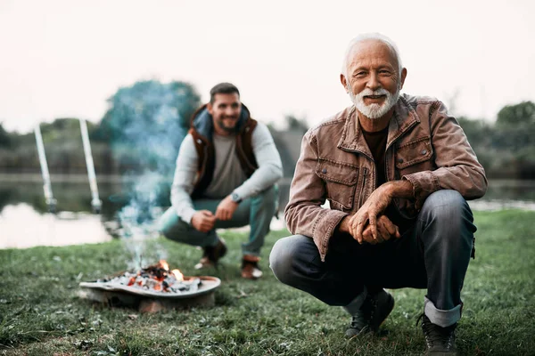 Happy mature man camping by the lake with is son and looking at camera.