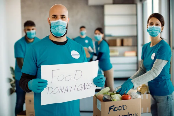 Man with face mask holding placard with \'food donations\' inscription while other volunteers are packing food in donation boxes.