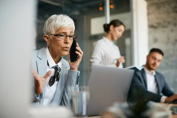 Mature female CEO communicating with someone over cell phone while working on laptop in the office.