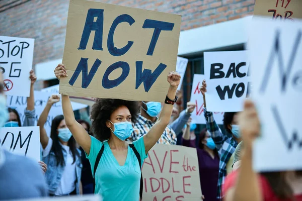 Displeased crowd of people with protective face mask on a protest during coronavirus pandemic.  Focus is on black woman carrying a banner with act now inscription.