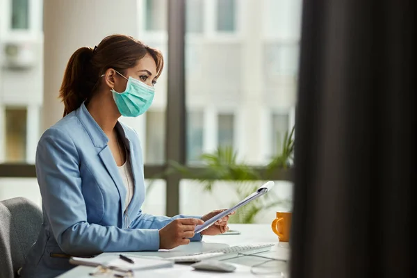 Pensive female entrepreneur with face mask working on business reports in the office. Copy space.