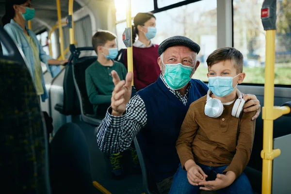 Happy senior man talking to his grandson while commuting by bus and wearing face masks dur to COVID-19 pandemic.
