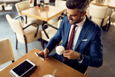 High angle view of happy businessman drinking coffee and texting on mobile phone while relaxing in a cafe.