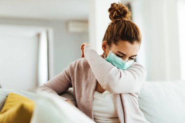Young woman with protective face mask sneezing into elbow while being at home. 