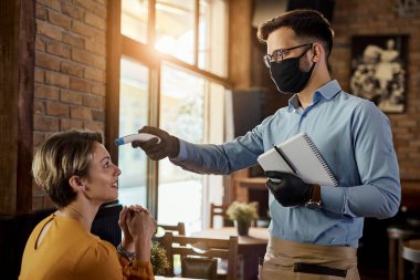 Young waiter wearing protective face mask and using infrared thermometer while measuring guest's temperature in a cafe.