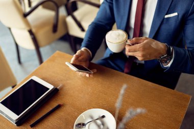 Unrecognizable businessman drinking coffee while text messaging on smart phone and relaxing in a cafe. 