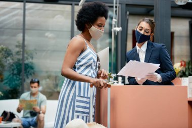 African American woman and receptionist wearing protective face masks going through registration documents during hotel check-in.
