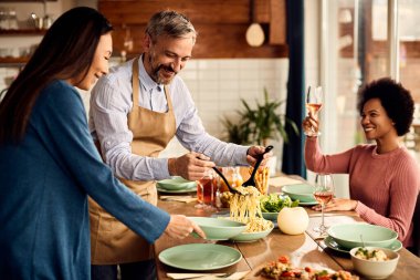 Happy man serving pasta to his friends while eating lunch in dining room.