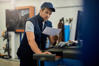 Otomobil tamirhanesinde kağıt işleriyle uğraşırken bilgisayar kullanan orta yaşlı bir tamirci.. 