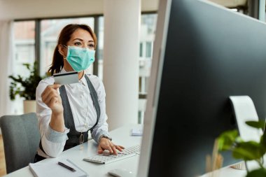 Female entrepreneur wearing face mask while using credit card and shopping on the internet in the office. 