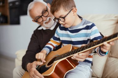 Happy senior man teaching his grandson to play acoustic guitar at home. Focus is on boy. 