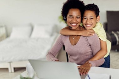 Happy black mother working on laptop while son is embracing her at home. They are looking at camera. 