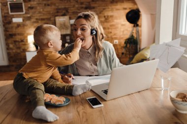 Mid adult single mother working at home while son is feeding her with a tangerine. 