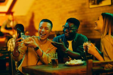 Group of happy friends having fun while drinking beer and using mobile phone in a bar. 