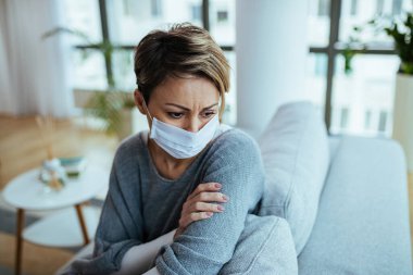 Distraught woman sitting on the sofa while wearing protective face mask and thinking during COVI-19 epidemic. 