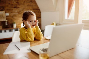 Distraught small girl sitting at the table and using computer at home. 