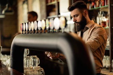 Young barista pouring beer from beer tap while working in a pub. 