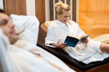 Happy woman relaxing with her husband at the spa and reading a book. 