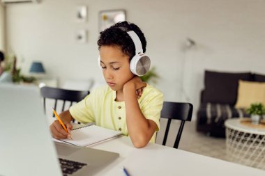 Black boy writing in notebook while using laptop and e-learning at home. 