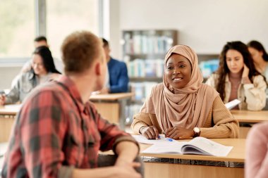 Happy African American female student in hijab communicating with her friend during a class in the classroom.