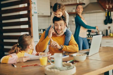 Working father talking on the phone while daughters are demanding his attention at home. Mother is preparing a meal in the background. 