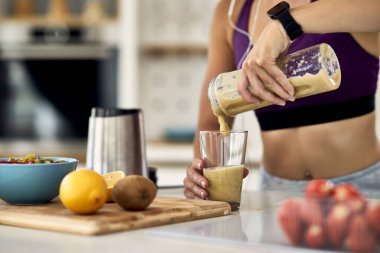 Close-up of sportswoman pouring fruit smoothie in a glass in the kitchen. 
