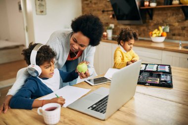 Happy African American mother giving a healthy snack to her son who is using laptop and learning from home. 