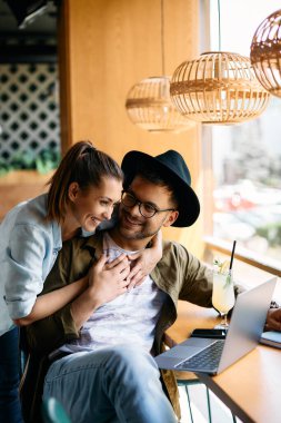 Young happy woman embracing her boyfriend who is using laptop and e-learning in a cafe.