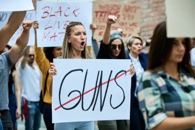 Young Caucasian woman shouting while protesting against use of weapons with crowd of people on the streets. 