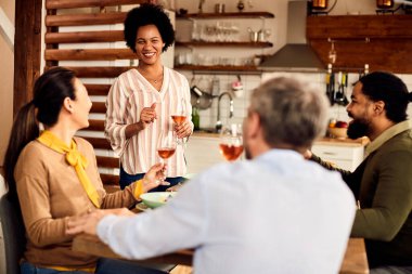 Multi-ethnic group of happy friends having lunch and drinking wine in dining room. Focus is o African American woman holding a toast. 